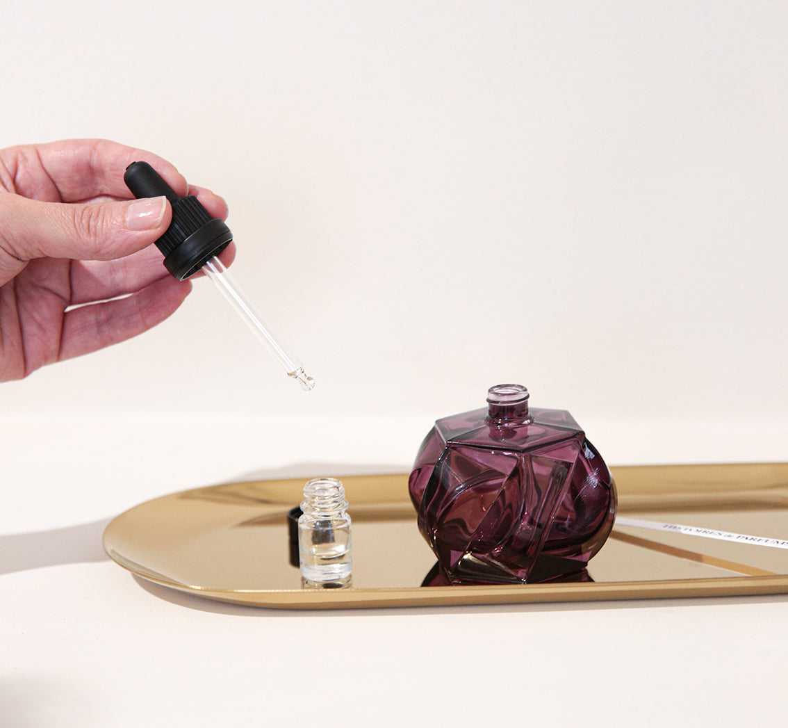 Hand using a dropper to transfer liquid from a purple bottle onto a white surface.